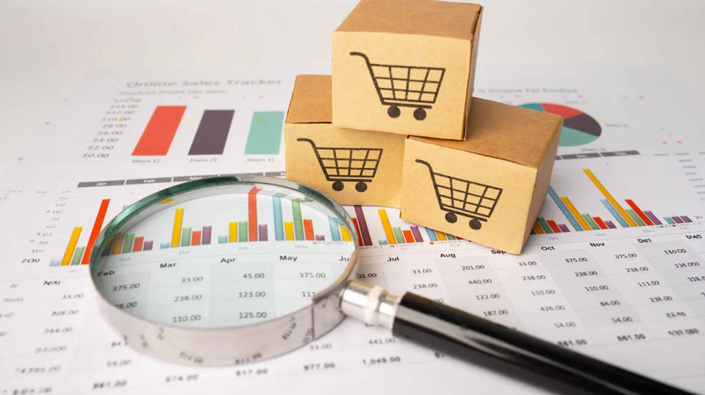 Cardboard boxes with shopping cart symbols, a magnifying glass, and sales charts illustrating retail sales analysis and business performance.